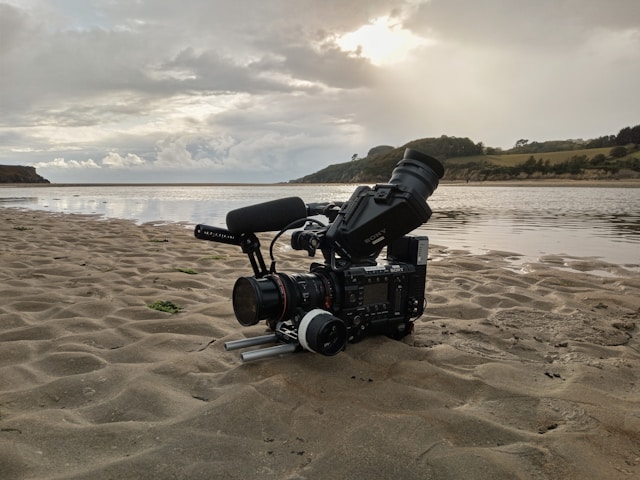 A video camera positioned in the foreground on sand with water behind it, illuminated by sun peaking out from a cloudy sky.